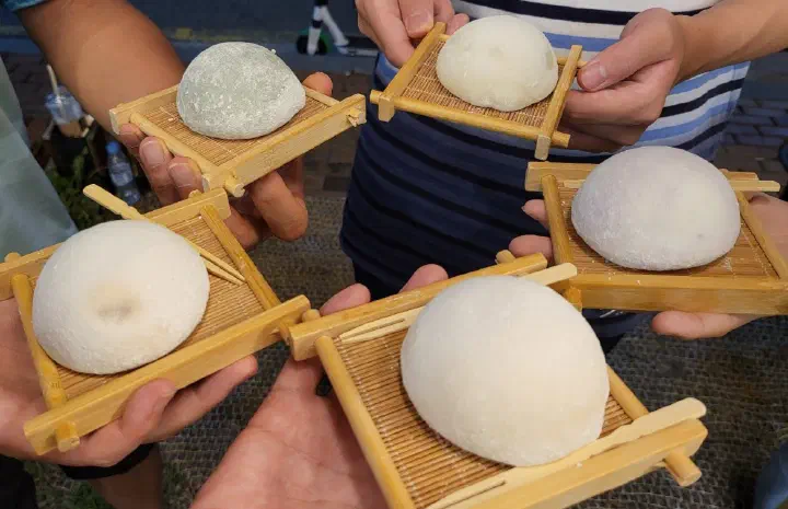 A group of friends each holding a jella tteok on a bamboo tray outside the shop in Haeundae