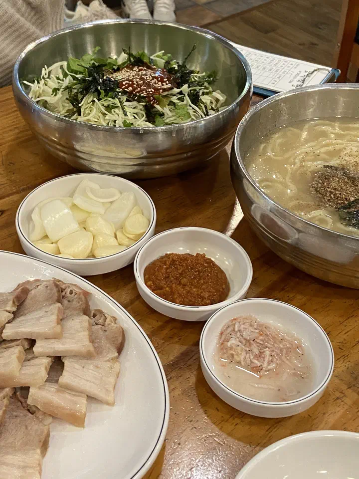 Bibimguksu and kalguksu on a wooden table with garlic, onion and salted shrimp side dishes