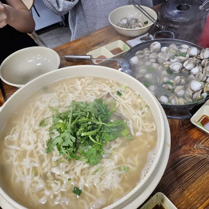 OC Kalguksu son-kalguksu, a big bowl of hand-cut clam noodle soup topped with crown daisy, next to the mulchong clam pot