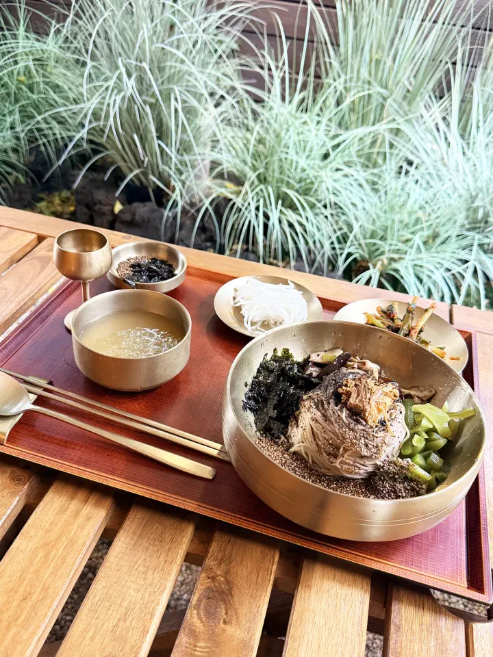 A makguksu set laid out on a wooden terrace table at Apgujeong Sanghoe beside ornamental grasses