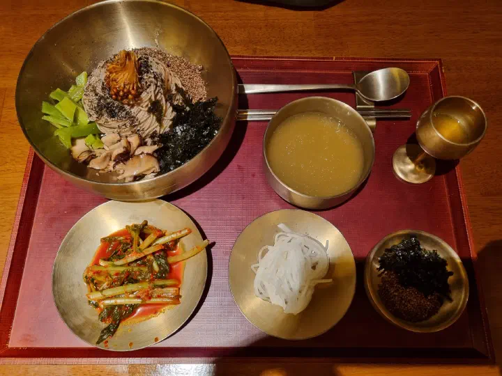 Deulgireum makguksu at Apgujeong Sanghoe served in a brass bowl with chilled broth, yeolmu kimchi, shredded radish and seaweed on a red tray