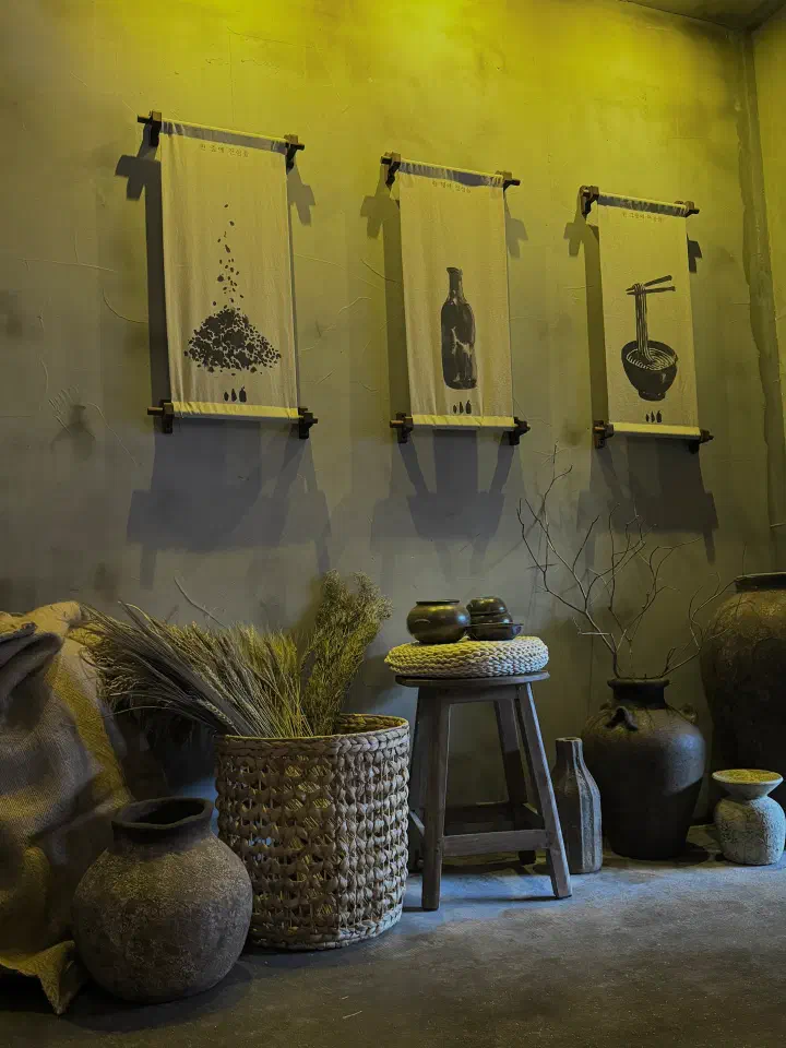 Fabric scrolls at Apgujeong Sanghoe illustrating perilla seed, an oil bottle and a bowl of noodles, above clay jars and dried grasses