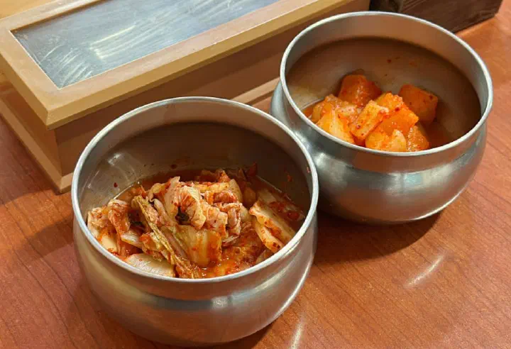 Cabbage kimchi and cubed radish kkakdugi in stainless steel bowls at Geumcheon Kalguksu