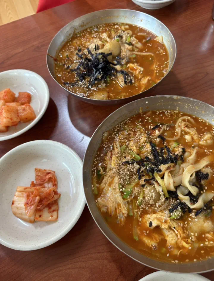Two bowls of jang kalguksu and ongsimi topped with dried seaweed, served with kimchi