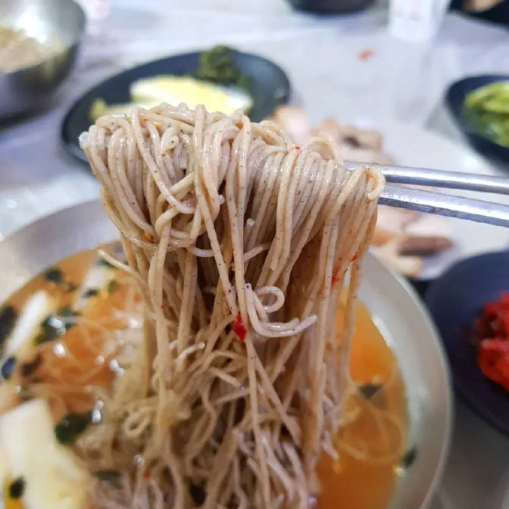 Chopsticks lifting Baekchon Makguksu buckwheat noodles over dongchimi broth