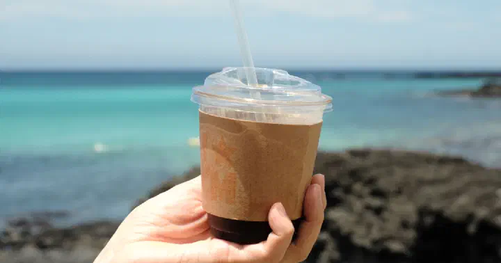 Hand holding a Cookiemeow cold brew coffee against the turquoise water of Hyeopjae Beach