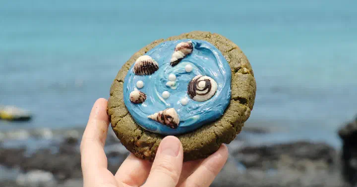 Jeju Badang matcha cookie topped with blue chocolate waves and shells, held in front of the Jeju sea