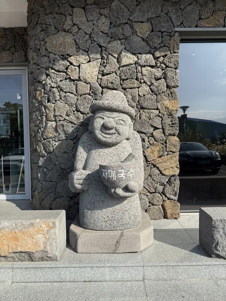 Jeju dol hareubang statue holding a bowl of Jamae Guksu noodles at the restaurant entrance photo spot