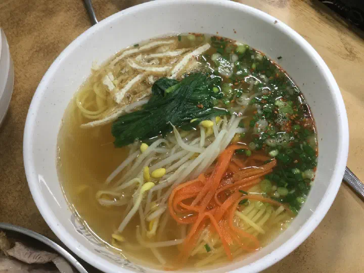 Bowl of clear warm broth noodles served alongside the bibim-guksu at Jamae Guksu, with bean sprouts, carrot, spinach and fried tofu