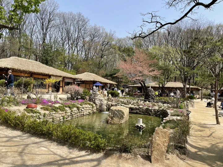 Guests waiting in the Mokhyangwon courtyard by the pond and thatched dining halls on a spring afternoon