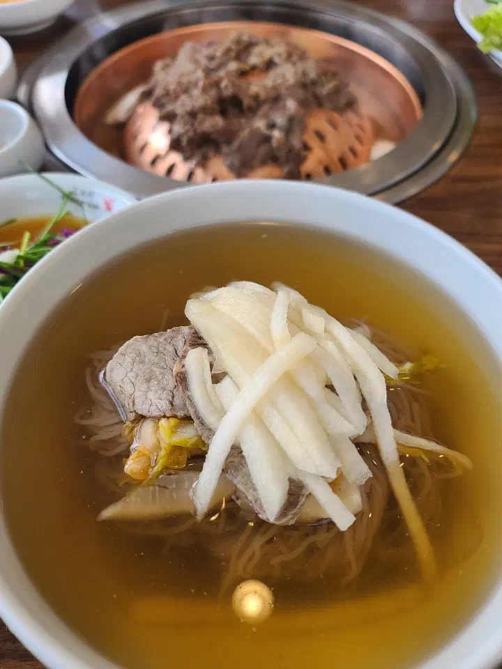 Woo Lae Oak Pyongyang naengmyeon in front with Hanwoo bulgogi grilling on the table behind it