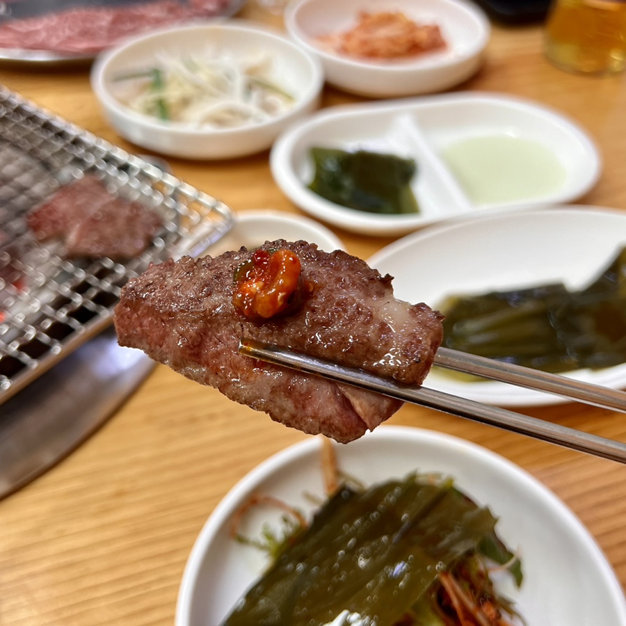 Grilled Hanwoo galbisal picked up with chopsticks, side dishes in the background