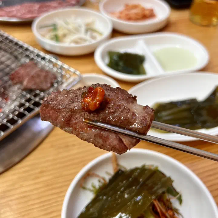 Grilled Hanwoo galbisal picked up with chopsticks, side dishes in the background