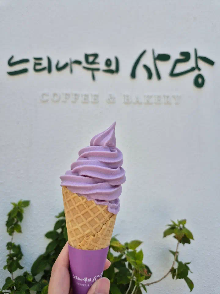 Lavender soft-serve ice cream cone held up in front of the Neutinamu-ui Sarang Coffee & Bakery sign
