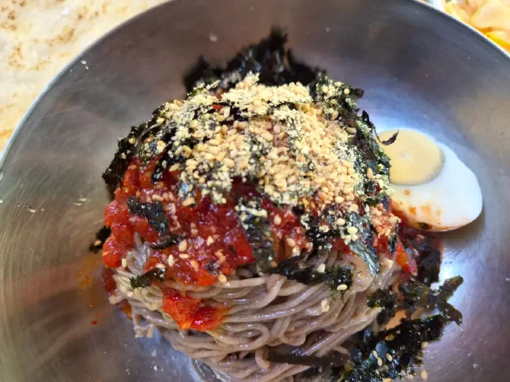 Cold buckwheat noodles in a stainless bowl topped with seaweed flakes, red sauce and half a boiled egg