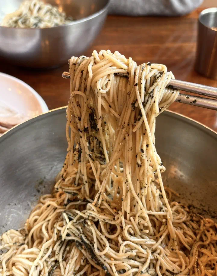 Lifting buckwheat noodles from a bowl of Gogiri Makguksu original perilla oil makguksu with chopsticks