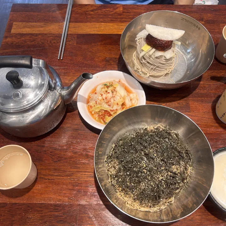 Kettle of chilled broth on the table beside a bowl of perilla oil makguksu at Gogiri Makguksu